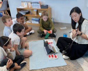 Montessori Classroom with Teacher/ Guide teaching the children in a semi circle on the floor.
